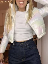 Campera puffer corta, acolchada, con acabado iridiscente que refleja tonos pastel. Posee cuello alto y cierre frontal con cremallera.