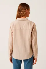 Blusa color beige claro, de corte holgado, con cuello camisero, escote en V y cierre parcial con botones. Presenta mangas largas con puños abotonados y dos bolsillos de parche en el pecho.