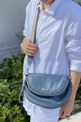 Bandolera celeste de cuero con forma de media luna, cierre superior con cremallera y tachas plateadas. Tiene correa de cuero celeste y correa de cinta regulable con rayas beige, verde y gris.