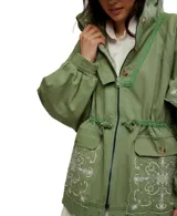 Chaqueta estilo parka o anorak de algodón color verde militar, con capucha y cordón ajustable en la cintura. Presenta parches de tela con bordados blancos de estilo paisley o floral en la parte delantera, mangas y capucha. El ruedo inferior tiene un borde festoneado con ojales.