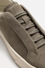 Zapatilla deportiva de hombre color gris topo, fabricada en piel con acabado serraje. Presenta cordonera elástica oculta y suela de goma de bajo volumen.