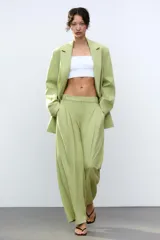 Pantalón verde claro de tiro alto con trabillas y botón en la cintura. Tiene pliegues, bolsillos laterales y falsos bolsillos de vivo en la espalda. El cierre frontal es con cremallera, botón interior y ganchos metálicos.