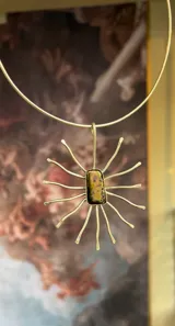 Gargantilla tipo collar rígido de metal dorado, con un dije colgante que simula un sol o estrella con un cristal rectangular central de tonos ámbar y negro.