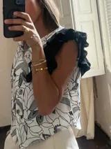 Chaleco blanco con estampado de hojas en negro, cuello camisero, cierre frontal con botones y mangas cortas con volados negros.