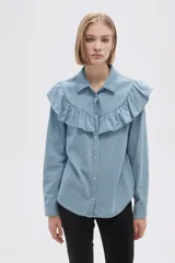 Camisa de jean azul con cuello clásico, cierre frontal con botones y mangas largas. Presenta volados en el canesú delantero y trasero.