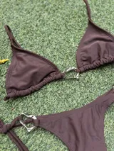 Conjunto de bikini color marrón chocolate, con corpiño triangular y bombacha colaless con tiras laterales para atar. Ambas piezas cuentan con argollas metálicas decorativas en el centro y los laterales.