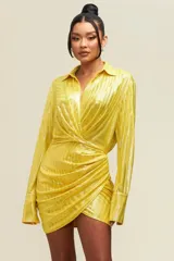 Minivestido camisero amarillo con textura brillante, corte cruzado y mangas largas con puños abotonados.
