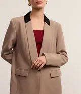 Blazer de tejido plano color beige con estructura de sastrería, cuello smoking con contraste negro en la nuca, botón único para cierre y solapas delanteras que simulan bolsillos.