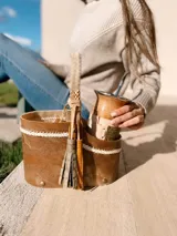 Bolso matero de cuero color marrón con divisiones internas para transportar termo, mate y yerba. Tiene manija trenzada y flecos decorativos.
