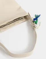 Cartera tipo saca color crudo, de cuerina, con asa corta regulable para el hombro y cierre superior. Incluye charms decorativos colgantes.