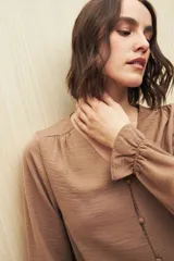 Camisa marrón de manga larga con escote en V y botones forrados al tono en el frente. Destaca por su textura sutil y por el detalle de frunces con volados en la parte inferior.