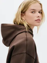 Buzo con capucha de color marrón oscuro, de corte holgado y mangas largas con costuras visibles. Se vislumbra un borde naranja debajo del buzo y un pantalón de jogging marrón con cordón blanco en la cintura.