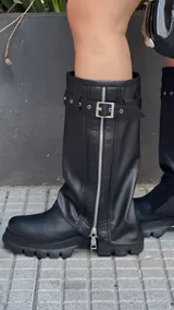 Botas de caña alta color negro, con suela track y cierre lateral con cremallera. Presentan una correa con hebilla alrededor de la caña.
