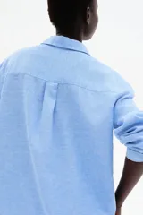 Camisa celeste de tejido liviano en mezcla de lino y viscosa, con cuello clásico, botones al frente, canesú de doble capa con pliegue de caja en la espalda, mangas largas con puños abotonados, bolsillo superior abierto y bajo redondeado.