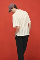 Camisa de manga corta color crema, confeccionada en tejido de punto jacquard con textura calada. Tiene cuello inglés, tapeta clásica con botones y bajo recto.