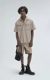 Bermuda estilo técnico color beige, confeccionada en microfibra liviana. Presenta cintura elástica con cinturón integrado de cinta negra con hebilla de plástico. Cuenta con múltiples bolsillos cargo, incluyendo uno lateral con panel de malla.