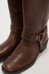 Bota de caña media color negro, estilo motociclista o cowboy, con puntera cuadrada y taco bajo. Presenta una tira ancha con hebillas metálicas alrededor del tobillo.