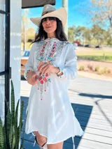 Vestido corto blanco con bordado floral multicolor en el frente, escote redondo con cierre de cremallera parcial y mangas largas.