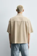 Camisa de denim de algodón, color beige claro, con corte relajado, cuello bowling y manga corta. Presenta cierre frontal con botones a presión y un pequeño bordado a tono en el pecho.