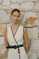Vestido midi de lino color beige, con escote cruzado en pico y sin mangas. Presenta vivos a contraste en negro y cinturón con hebilla forrada. Cierre frontal con botones.