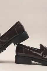 Mocasín negro de cuero vacuno con acabado charol, suela de goma y forro y plantilla de cuero vacuno.