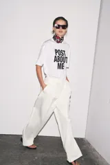Camiseta blanca de corte amplio, cuello redondo y mangas cortas. Estampado frontal con la frase "Post About Me" en letras negras.