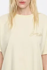 Camiseta color crudo de algodón, con cuello redondo y mangas cortas. Presenta logo bordado en color dorado en el frente.