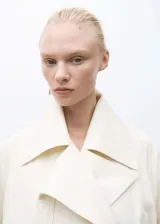 Abrigo largo color crema con cuello de solapa ancha, trabillas en los puños, cinturón desmontable y dos bolsillos laterales.