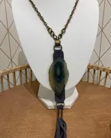 Collar con cadena de eslabones color bronce, dije de piedra natural con forma irregular en tonos verde y azul, y borla de flecos de gamuza gris.