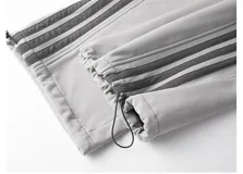 Pantalón deportivo gris claro, estilo cargo o jogging, con franjas negras verticales en los laterales de las piernas. Posee tobillos regulables y cintura ajustable.