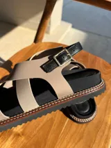 Sandalias bajas color beige con tiras cruzadas y hebilla al tobillo. Suela de goma negra con pespuntes blancos.