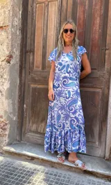 Vestido largo con estampado paisley en tonos azules y violetas sobre fondo blanco. Presenta escote en V, mangas cortas con volados y un ruedo con volado.