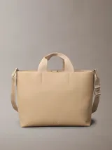 Cartera tote Calvin Klein color beige, de piel sintética granulada, con doble asa corta y correa larga ajustable con logo jacquard. Incluye cierre de correa magnético y bolsillos interiores.
