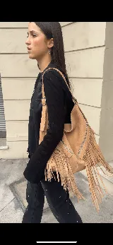 Bolso tipo hobo color tostado, hecho de cuero con flecos largos en el frente y dos bolsillos con cierre. Tiene una manija regulable con hebillas metálicas.