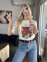 Remera blanca de cuello redondo y manga corta con estampado de paisaje costero en tonos naranja, rojo, verde y azul.