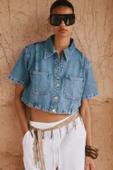 Camisa de denim celeste de corte crop, con cuello solapa, escote en pico, manga corta y dos bolsillos delanteros con botón. Cierre frontal con botones metálicos.