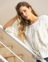 Blusa color crema con cuello redondo, abertura central con botones y mangas 3/4 con puño fruncido. Presenta detalles calados y bordados en mangas, delantera y espalda. La cintura se ajusta con un lazo.