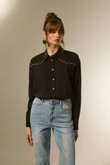 Camisa holgada de manga larga, color negro, con cuello camisa y botones frontales. Presenta apliques de tachas plateadas en los hombros y a lo largo de la tapeta frontal.