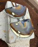 Collar con colgante de dos piedras naturales irregulares engarzadas en metal plateado y dorado, suspendidas de un cordón de cuero marrón con detalle de nudo corredizo. Las piedras tienen vetas en tonos naranja, marrón y blanco.