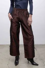 Pantalón de tafeta tornasolada color borgoña, de corte recto con cintura elastizada, bolsillos frontales con cierre plateado, y cordones ajustables en las pantorrillas para fruncir el bajo.