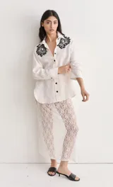 Camisa blanca de lino con bordados florales en los hombros, de manga larga y corte recto con abertura en los laterales. Cierre frontal con botones negros.