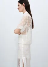 Camisa blanca semitransparente con bordado de flores, cuello camisero, manga corta y cierre frontal con botones.