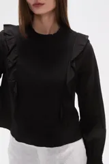 Blusa de algodón color negro con cuello a la base y detalle de volados en el frente. Posee mangas largas con puños elásticos y un panel frontal con textura fruncida.