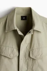 Sobrecamisa color arena de lona gruesa de algodón, corte recto, con cuello clásico, cierre frontal con botones a presión ocultos, canesú en la espalda, dos bolsillos superiores con solapa y botón a presión, bolsillos laterales discretos, mangas largas con puños abotonados y forro interno.