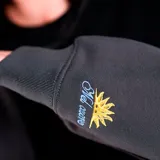 Sudadera tipo canguro negra con capucha y bolsillo canguro frontal. Presenta un diseño de doble manga, donde las mangas interiores son de color gris oscuro. Tiene un pequeño logo bordado en blanco en el pecho izquierdo.