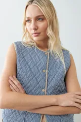 Chaleco capitoneado reversible a rayas azules y blancas con ribete color crema. Tiene cuello redondo, cierre con botones a presión y bolsillos de parche. El reverso es liso color tostado.