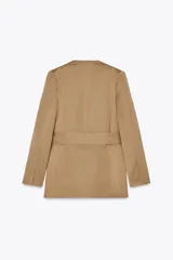 Blazer color beige con escote en pico sin solapa, manga larga con hombreras, bolsillos delanteros con solapa, cierre frontal cruzado con botones forrados a tono y cinturón del mismo tejido.