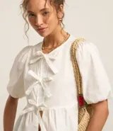 Blusa blanca de tricolina con cuello redondo, manga corta abullonada, cierre frontal con lazos y volado en la cintura.