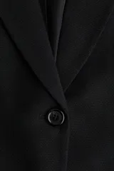 Blazer negro de corte largo y botonadura sencilla con un solo botón. Presenta solapas de muesca, bolsillos delanteros ribeteados y mangas tres cuartos con efecto remangado o fruncido.