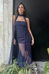 Vestido largo de color azul marino con base de crepé asimétrica y sobrecapa de microtul translúcido. Presenta un diseño de escote recto sin breteles y falda con transparencia. Incluye un accesorio de tela a juego versátil para usar como chal o lazo al cuello.